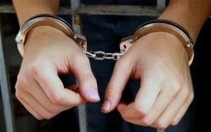 Două mâini de persoană adultă sunt prinse în cătușe metalice în fața unui corp îmbrăcat în haine închise la culoare.