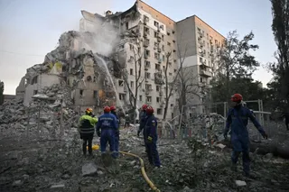 Mai mulți salvatori cu căști colorate stau lângă un bloc de apartamente partially distrus și un morman de dărâmături sub un cer deschis.