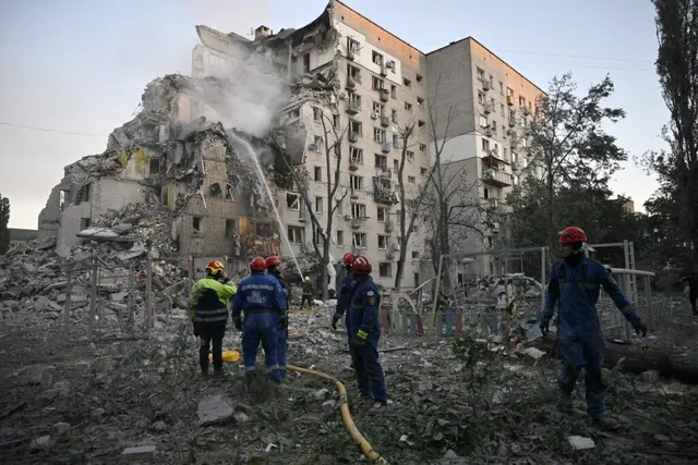 Mai mulți salvatori cu căști colorate stau lângă un bloc de apartamente partially distrus și un morman de dărâmături sub un cer deschis.
