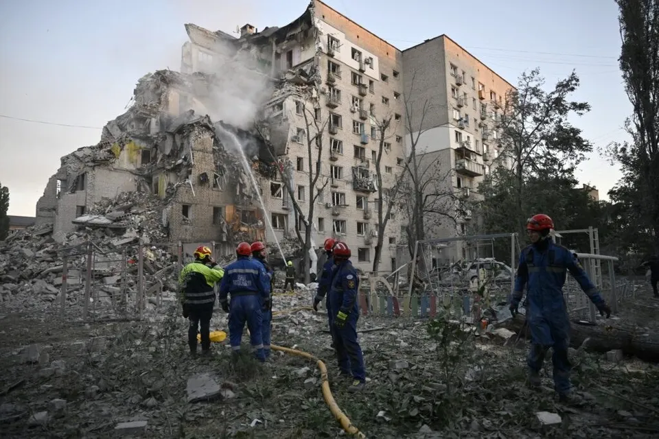 Mai mulți salvatori cu căști colorate stau lângă un bloc de apartamente partially distrus și un morman de dărâmături sub un cer deschis.