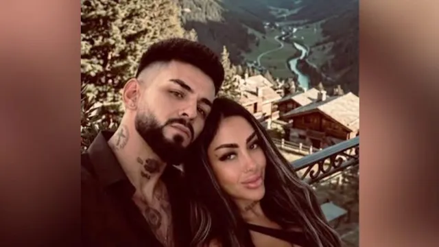 Un bărbat cu părul scurt și o femeie cu părul lung stau împreună în fața unui peisaj montan cu case și un râu.