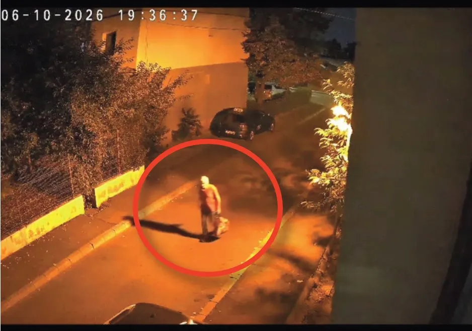 Un bărbat merge pe un trotuar lângă un gard, fiind încadrat într-un cerc roșu pe o imagine de supraveghere nocturnă.