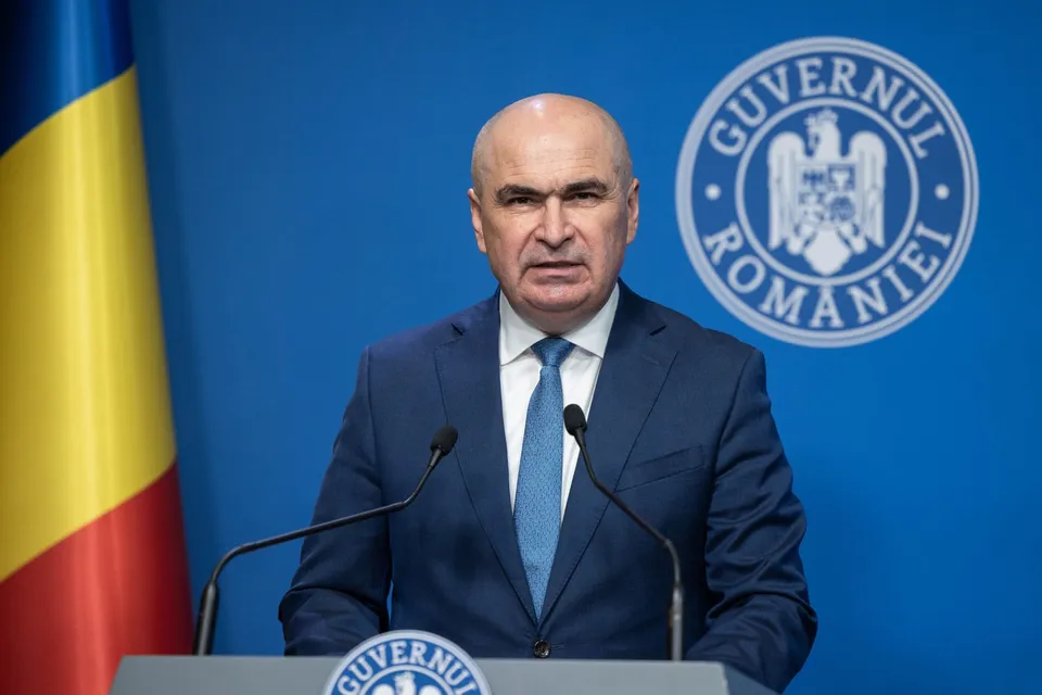 Un bărbat cu părul scurt și costum albastru stă în fața unui fundal albastru cu un emblemă circulară și un steag tricolor.