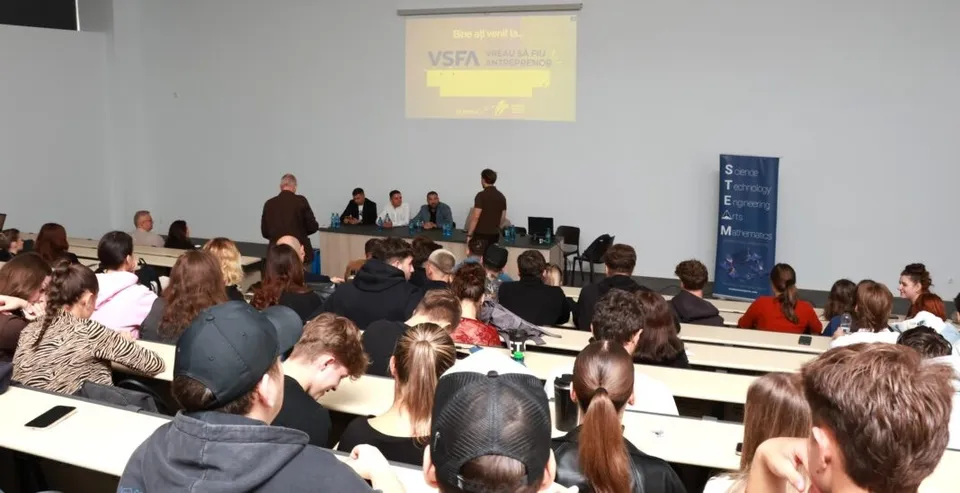 O sală de conferințe cu un grup de oameni așezați la mese lungi privind spre un ecran de proiecție și un grup de persoane de la o masă.