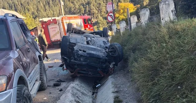 Un autoturism gri este răsturnat pe o parte într-un șanț, lângă un vehicul de serviciu roșu și o mașină de culoare închisă.