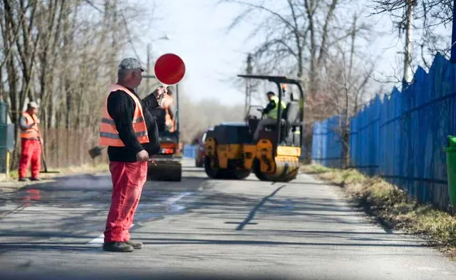 Un lucrător cu vestă reflectorizantă portocalie și pantaloni roșii stă pe un drum asfaltat lângă un cilindru compactor galben și alți muncitori.