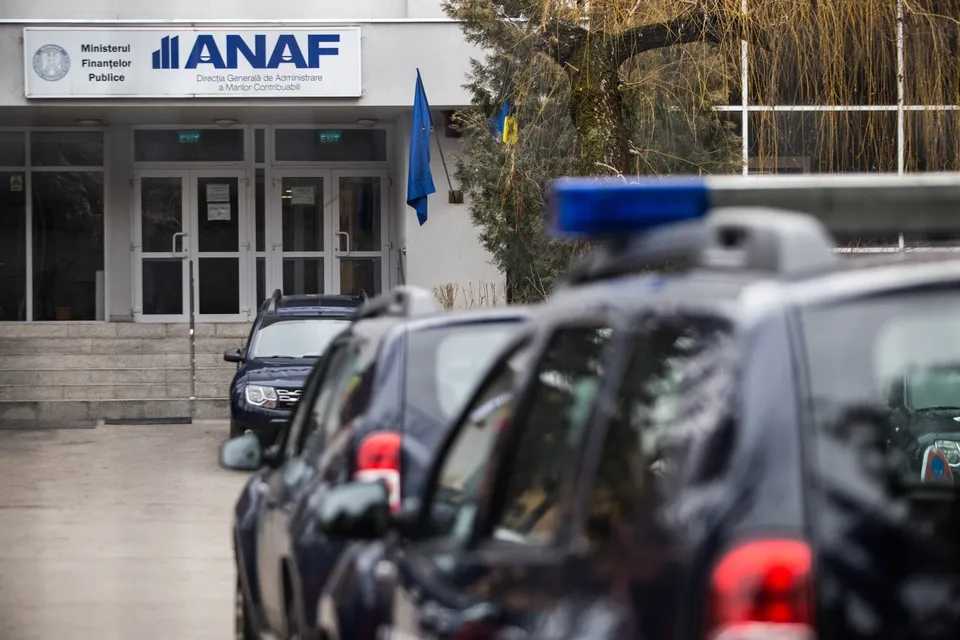 Mașini de poliție sunt parcate în prim-plan în fața unei clădiri pe care este afișat un panou cu inscripția ANAF.