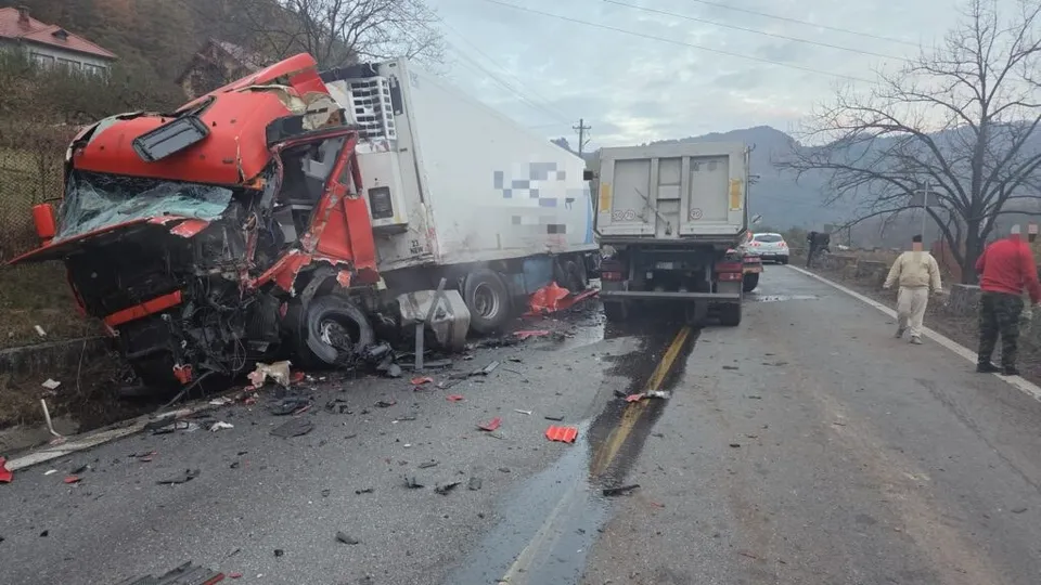 A red semi-truck is severely crushed on its left side, resting against a guardrail, with debris scattered across the road, while a silver truck and other vehicles are stopped nearby.