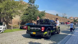 Un pickup negru cu autocolante verzi este parcat pe un drum pavat lângă un grup de oameni și alte vehicule sub un cer senin.