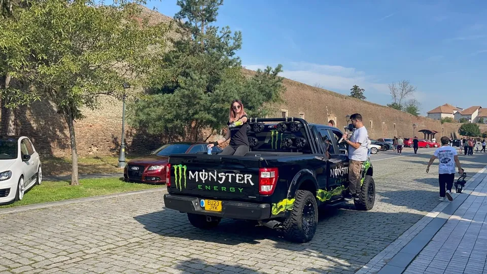 Un pickup negru cu autocolante verzi este parcat pe un drum pavat lângă un grup de oameni și alte vehicule sub un cer senin.