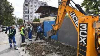 Un excavator galben sapă un morman de pămui lângă un grup de lucratori purtând veste reflectorizante și căști de protecție.