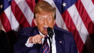 Un bărbat cu părul blond, purtând un costum albastru și o cravată cu dungi roșii, vorbește la un microfon în fața unor steaguri americane.