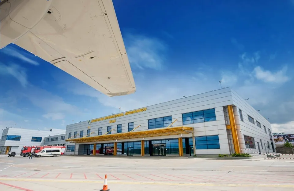 O aripă de avion se află în prim-plan deasupra unei clădiri de aeroport cu ferestre mari sub un cer albastru cu nori.