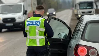Un polițist cu vestă reflectorizantă stă lângă portiera deschisă a unei mașini gri pe un drum cu alte vehicule în fundal.