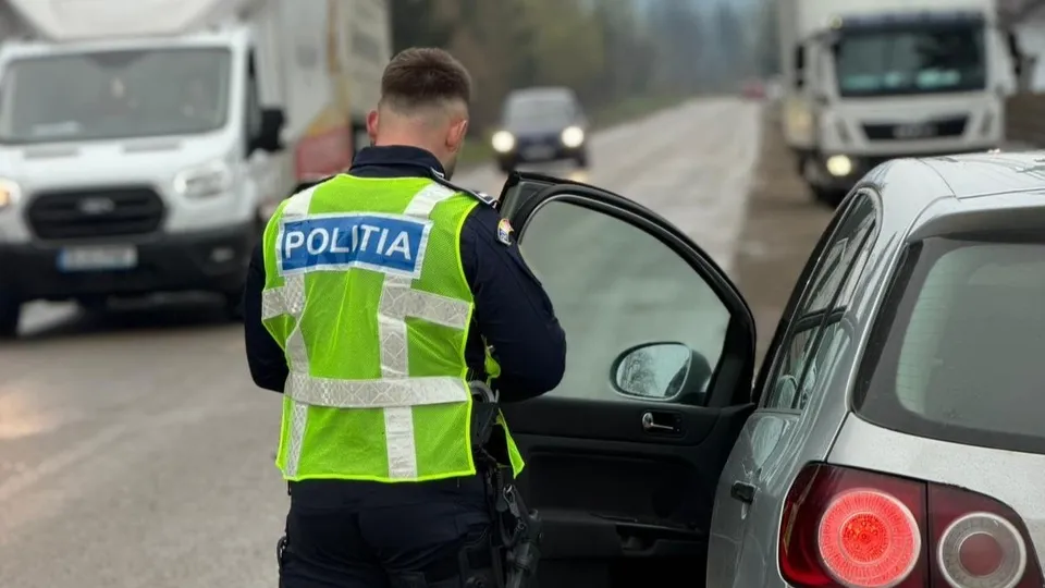 Un polițist cu vestă reflectorizantă stă lângă portiera deschisă a unei mașini gri pe un drum cu alte vehicule în fundal.