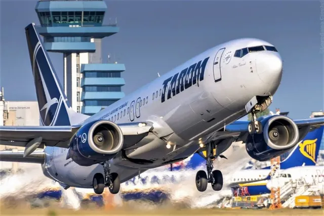 Un avion de pasageri gri cu inscripția TAROM decolă de pe o pistă, având două motoare suspendate sub aripi și trenul de aterizare extins.