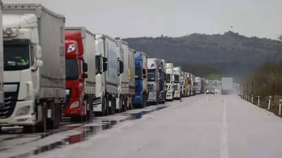 O coloană de camioane mari este parcată pe marginea unui drum asfaltat lângă un deal împădurit sub un cer înnorat.