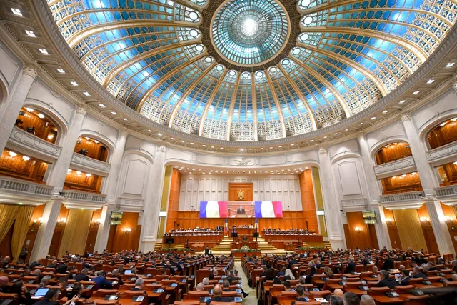 O sală de conferințe circulară cu un plafon de sticlă decorat, având mulți oameni așezați la mese de lemn dispuse în rânduri sub un palet de steaguri.
