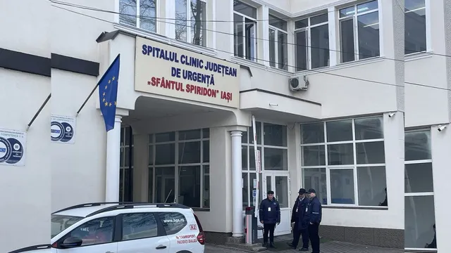 O clădire albă cu ferestre mari dispune de un afiș cu text și un steag albastru, în fața căreia stau trei persoane lângă o mașină albă.
