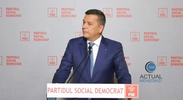 Un bărbat într-un costum albastru și cravată deschisă stă la un pupitru cu inscripția Partidul Social Democrat în fața unui fundal cu același logo repetat.