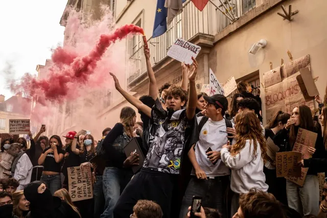 Un grup de tineri protestează într-o stradă, unul dintre ei ridicând un semn, în timp ce fum roz se ridică în fundal.