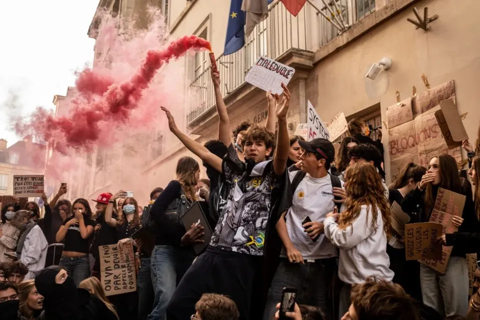 Un grup de tineri protestează într-o stradă, unul dintre ei ridicând un semn, în timp ce fum roz se ridică în fundal.