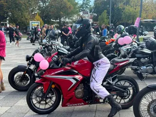 Un motociclist cu cască neagră și pantaloni albi stă pe o motocicletă roșie decorată cu baloane roz într-o zonă pietonală cu mulțime.