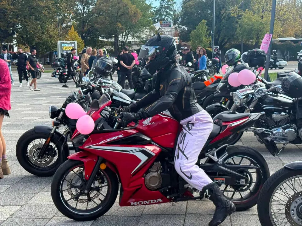 Un motociclist cu cască neagră și pantaloni albi stă pe o motocicletă roșie decorată cu baloane roz într-o zonă pietonală cu mulțime.