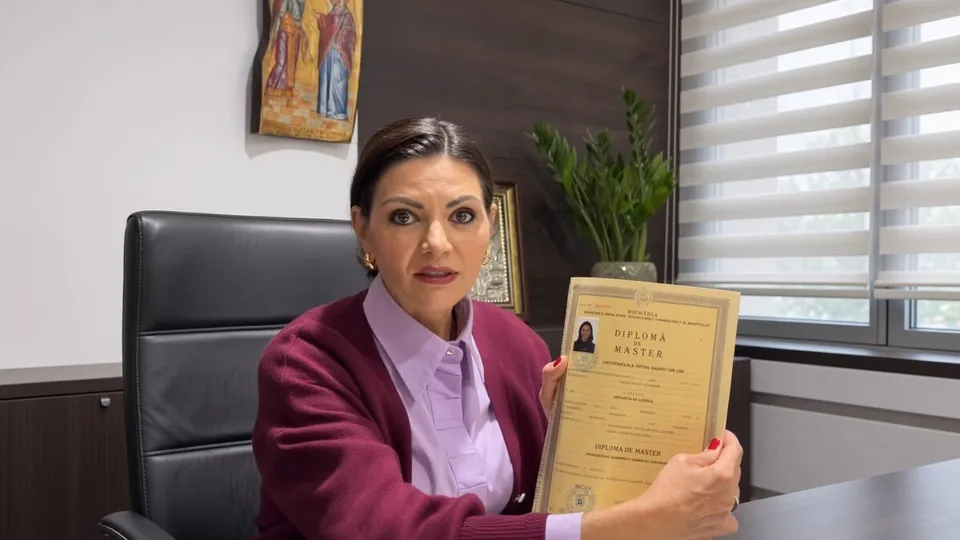 O femeie cu părul închis la culoare, purtând o sacou purpuriu și o cămașă lila, ține în mâini o diplomă de master într-un dosar galben.
