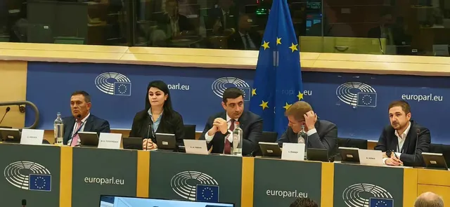 Cinci persoane stau la o masă lungă în fața unui panou albastru cu logo-uri și un steag european.