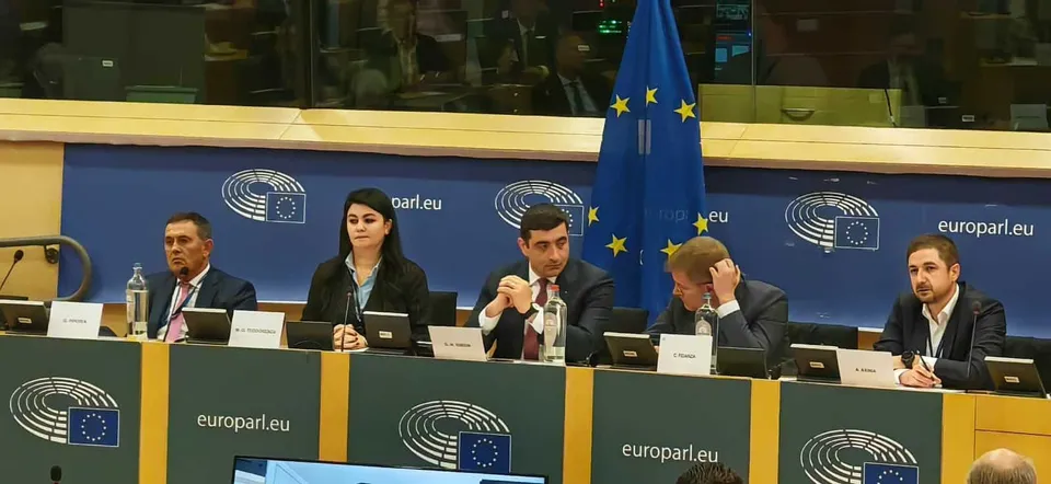 Cinci persoane stau la o masă lungă în fața unui panou albastru cu logo-uri și un steag european.