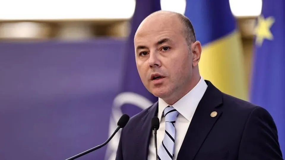 Un bărbat cu părul rare, purtând costum închis la culoare, cămașă albă și cravată albastră cu dungi albe, vorbește la microfoane.