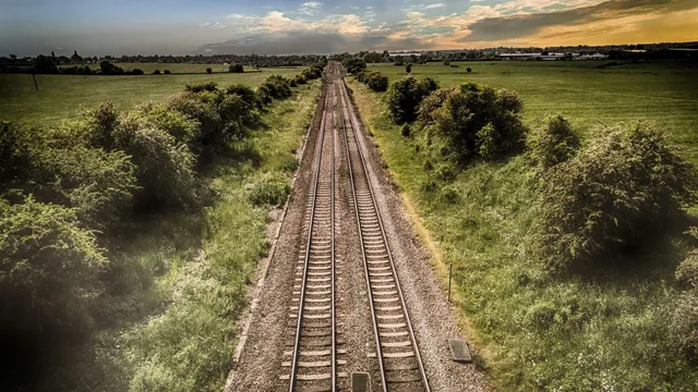 Două șine de tren întindute paralel prin vegetație și câmpuri deschise sub un cer cu nori la apus.