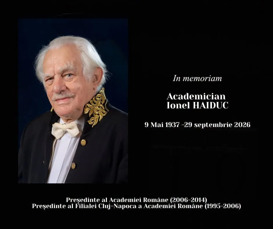 Un bărbat în vârstă cu părul alb și mustață poartă un frac negru, cămașă albă și papion alb pe un fundal închis.