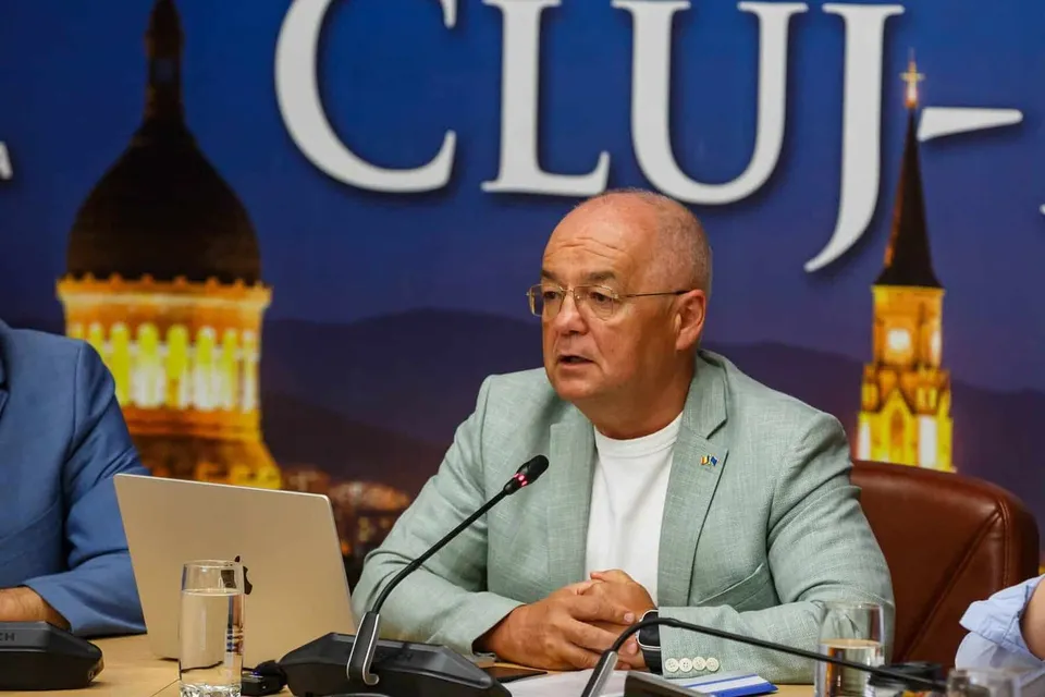 Un bărbat cu ochelari și sacou deschis stă la o masă lângă un laptop și microfoane.
