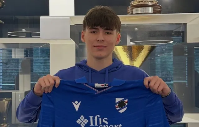 Un tânăr bărbat cu părul castaniu deschide un tricou de fotbal albastru cu logo-uri în fața unei vitrine cu trofee.