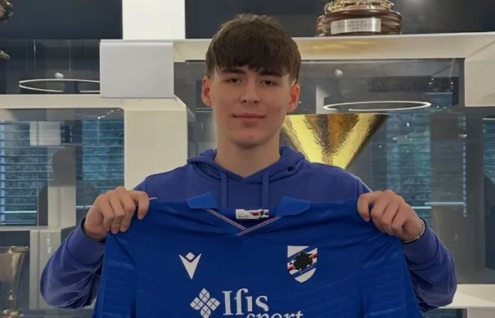Un tânăr bărbat cu părul castaniu deschide un tricou de fotbal albastru cu logo-uri în fața unei vitrine cu trofee.
