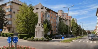 O cruce mare de piatră este amplasată pe un rond de flori în fața unor clădiri de apartamente și a unor copaci verzi.