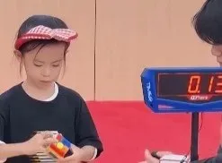 O copilă cu o bandană roz și albă ține un cub colorat în mâini, lângă un cronometru digital care afișează cifrele portocalii.