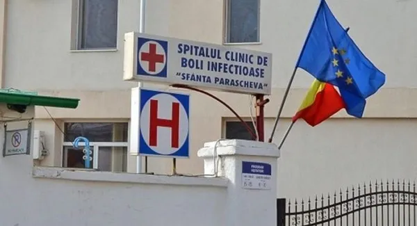 Un semn de spital cu o cruce roșie și un steag al Uniunii Europene sunt montate pe fațada unei clădiri albe.