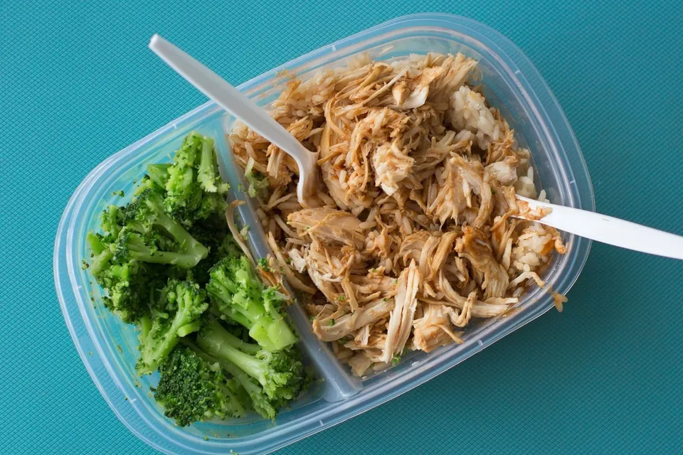 O caserolă transparentă cu două compartimente, unul cu broccoli și celălalt cu carne mărunțită, pe o suprafață albastră texturată.