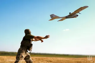 Un bărbat cu mască și vestă tactică aruncă un aparat zburător mic spre cerul senin deasupra unui câmp uscat.