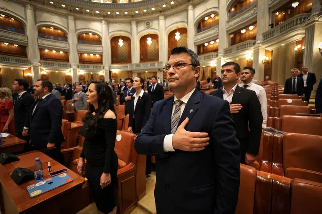 Un bărbat cu ochelari și costum în prim-plan stă în picioare cu mâna pe piept într-o sală de ședințe cu scaune de piele maro.