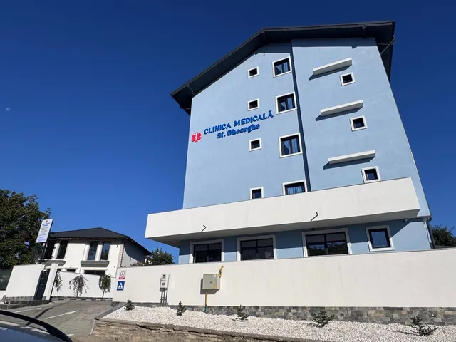 O clădire albastră cu ferestre albe este situată sub un cer senin, lângă un gard alb și un pavaj de pietriș.