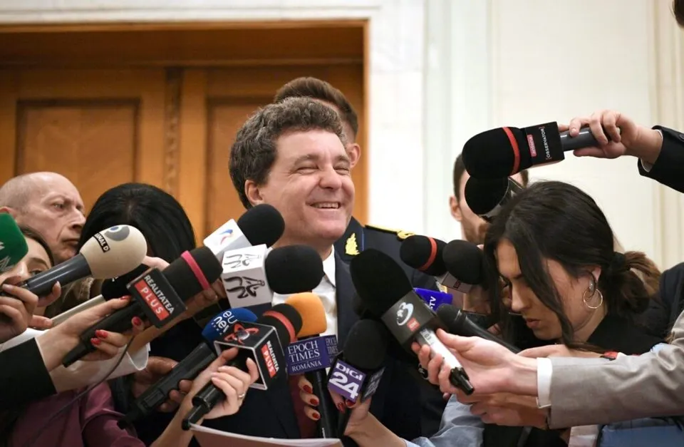 Un bărbat zâmbește în timp ce mai mulți oameni țin microfoane cu diverse logo-uri în fața lui.