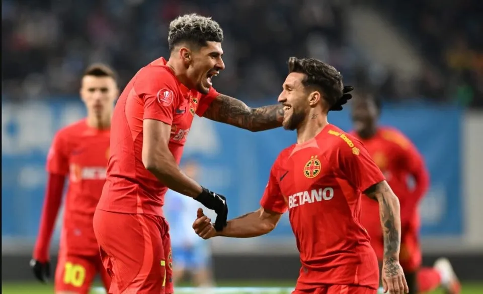 Doi jucători de fotbal în echipamente roșii sărbătoresc pe un teren de sport, în timp ce alți doi jucători sunt vizibili în fundal.