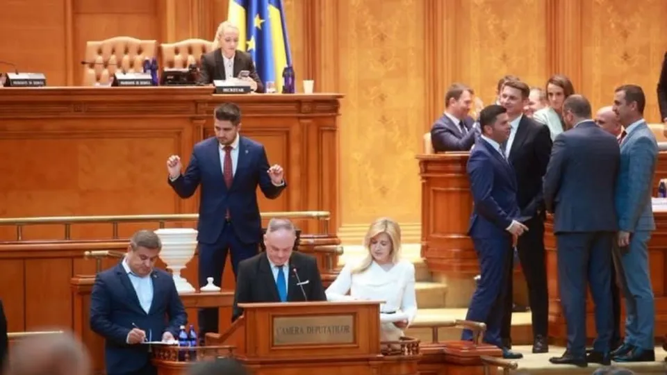 Un grup de bărbați în costume de culoare închisă stau în picioare într-o încăpere cu panouri de lemn, în timp ce un bărbat stă în picioare vorbind.