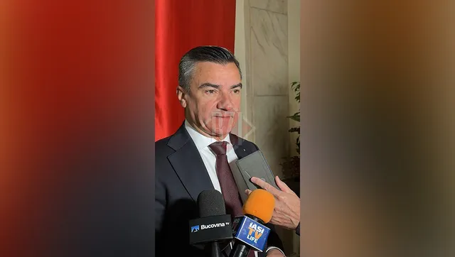 Un bărbat într-un costum închis la culoare și cravată roșie stă în fața mai multor microfoane de presă.