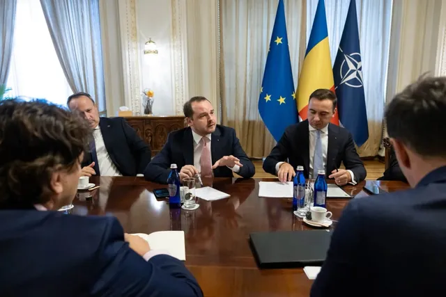 Cinci bărbați în costume purtând cămăși și cravate stau la o masă de lemn într-o încăpere cu steaguri în fundal.