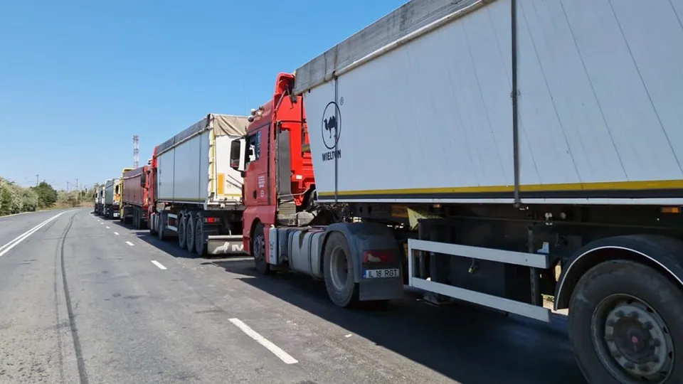 Mai multe camioane cu remorci de marfă sunt parcate în lanț pe marginea unei autostrăzi sub un cer senin.